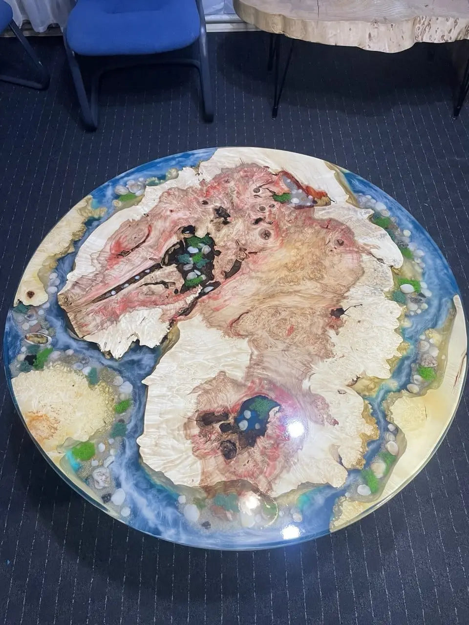 Artistieke ronde salontafel van massief hout met contrasterende houtnerven, helderblauwe epoxyhars rivier, ingelegd met echt mos en schelpen. Uniek design, geïnspireerd op natuur en kust, perfect voor moderne en luxe interieurs.