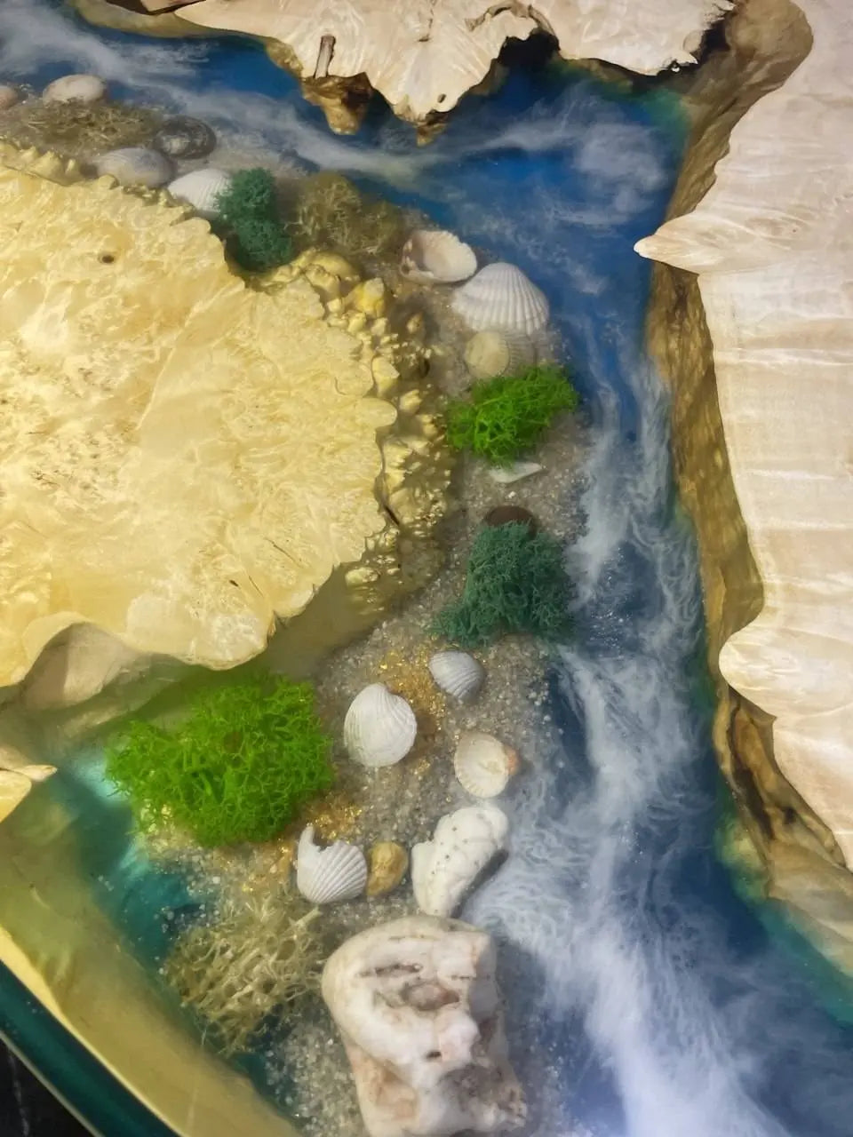 Een levendige epoxyhars salontafel met een blauwe rivier die stroomt tussen lichte houten oevers. Ingelegde schelpen, groen mos en ruwe stenen vormen een kustlandschap.