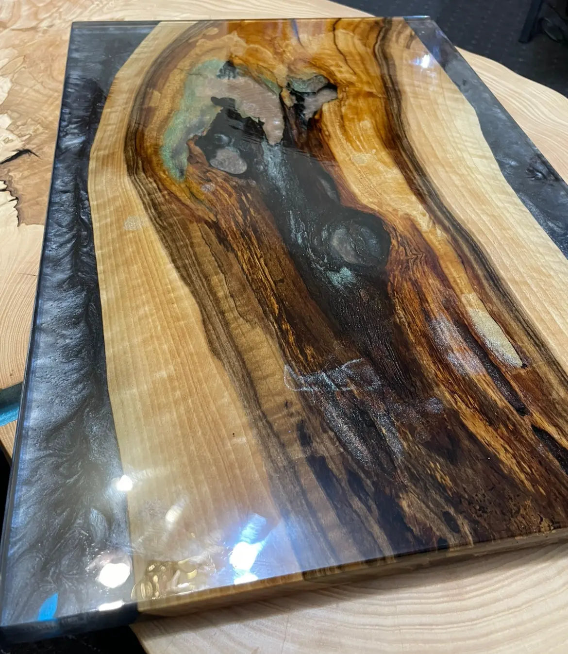 Handgemaakte snijplank van epoxyhars met inleg van Black Pearl River, tinten esdoorn en olijfhout met een levendige rand en een artistieke hoogglansafwerking. Ideaal voor moderne interieurs, woonkamers en luxe kantoorruimtes.