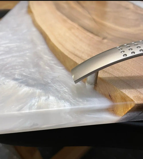 Close-up van een metalen handvat bevestigd aan de rand van een houten oppervlak, met daaronder een glanzend wit laken. De toon is industrieel en eigentijds.