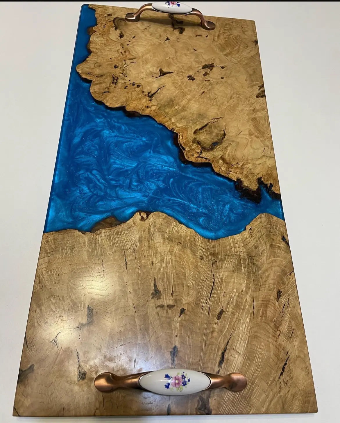 Een rechthoekige serveerschaal met een natuurlijk houtnerfpatroon en een levendige blauwe rivier van hars, met keramische handgrepen met bloemenmotief, op een wit oppervlak.