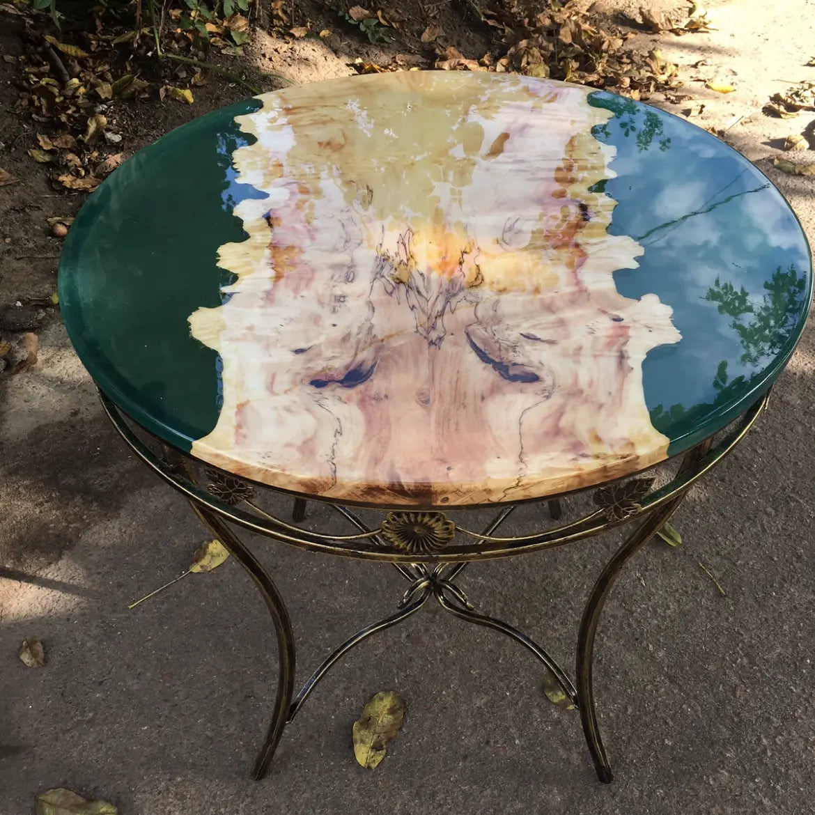 Stijlvolle tafel met een groen rond glazen blad en een houten onderstel met ingewikkelde patronen en een goudkleurig metalen frame, voor buiten.