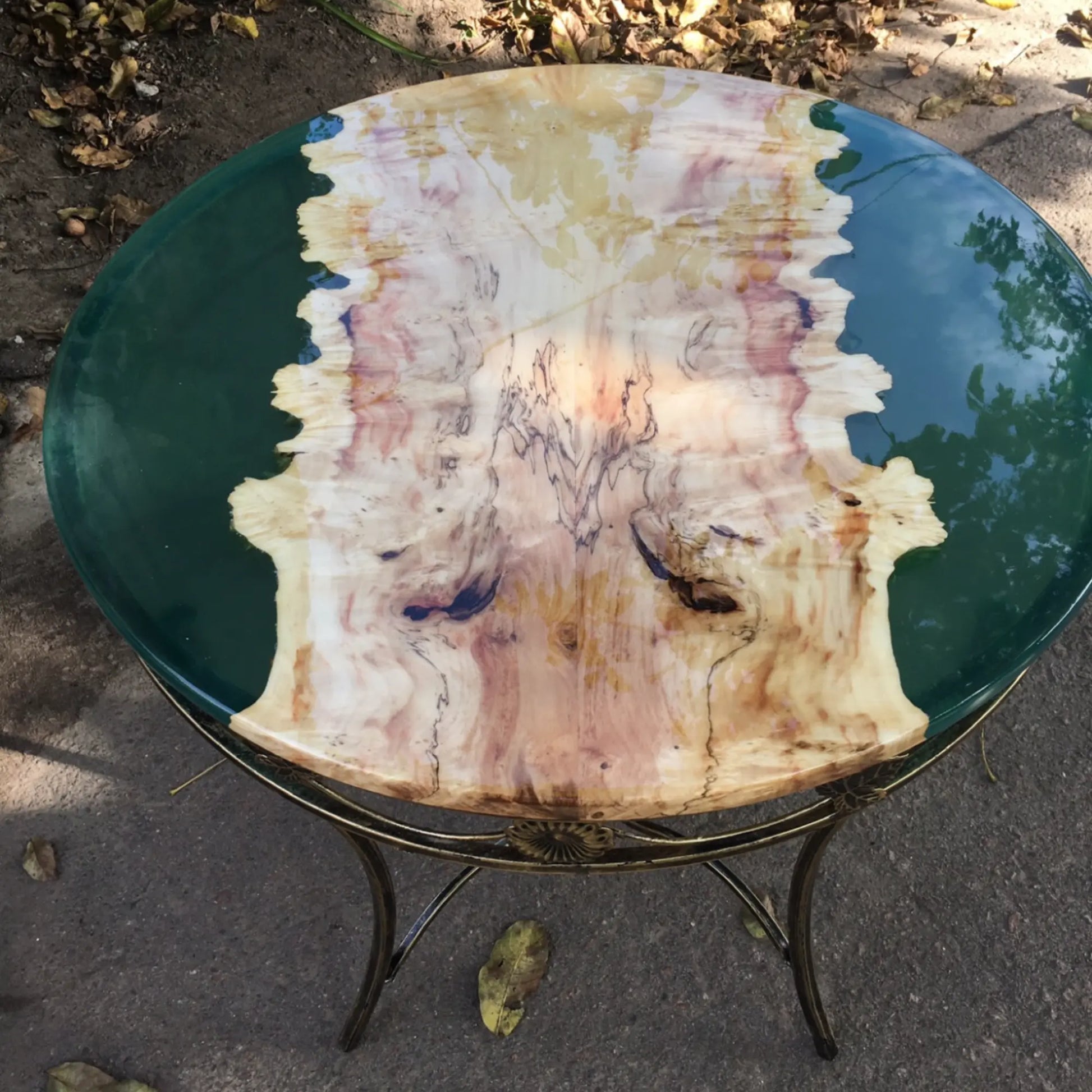 Design buitentafel met groen rond glazen blad, decoratief houten onderstel en goudkleurig metalen frame – luxe tuintafel voor moderne buitenruimtes.