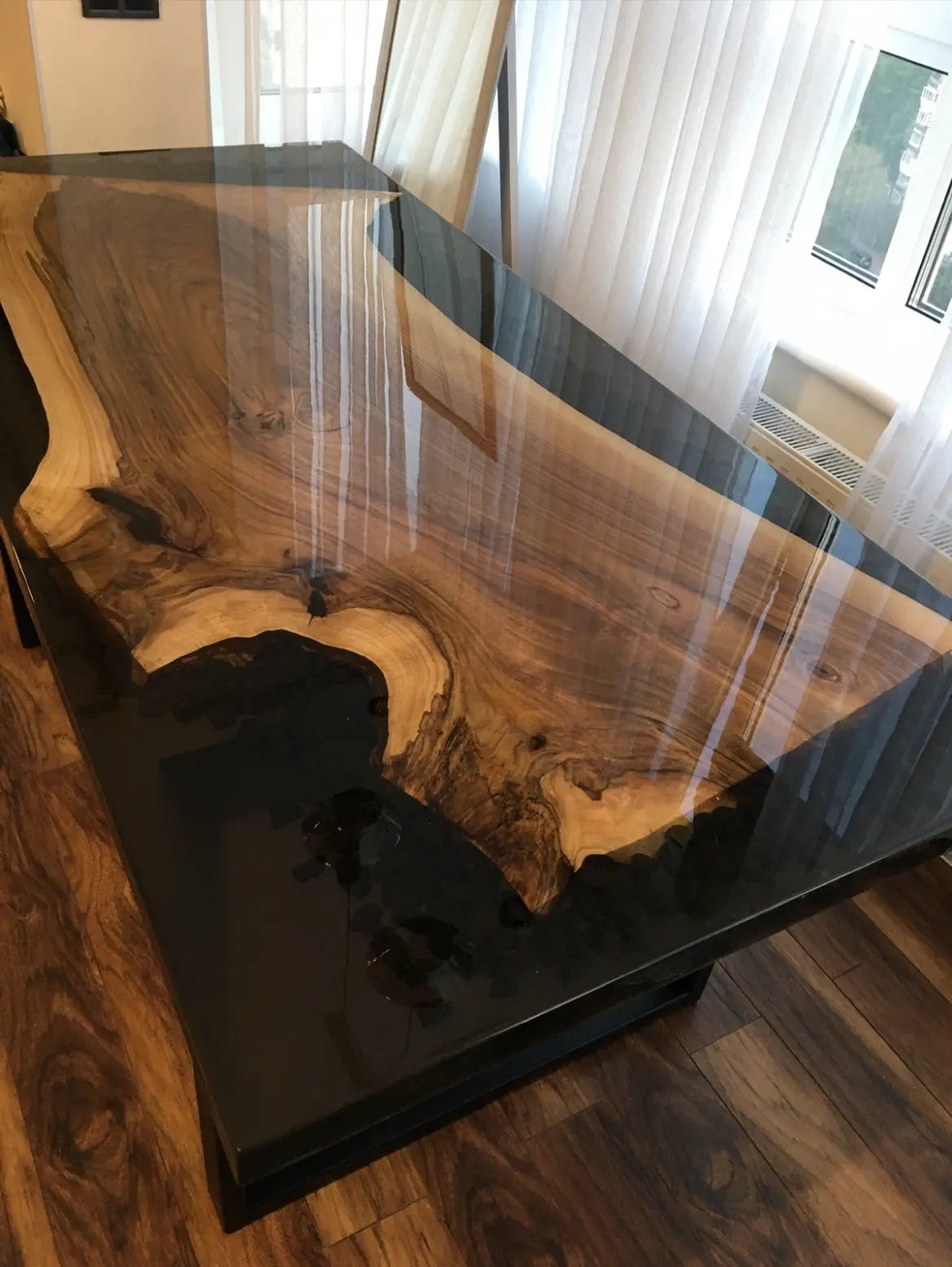 Een close-up van een houten tafel met een glanzend oppervlak, met natuurlijke houtnerfpatronen en een harsafwerking. De tafel staat in een goed verlichte kamer met grote ramen en een houten vloer.