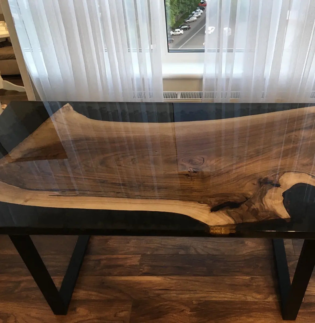 A close-up view of a wooden table with a glossy surface, showcasing natural wood grain patterns and a resin finish. The table is positioned in a well-lit room with large windows and wooden flooring.