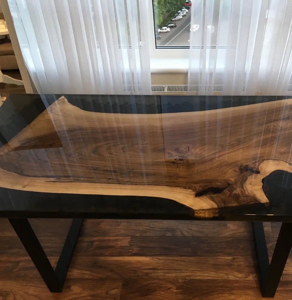 A close-up view of a wooden table with a glossy surface, showcasing natural wood grain patterns and a resin finish. The table is positioned in a well-lit room with large windows and wooden flooring.