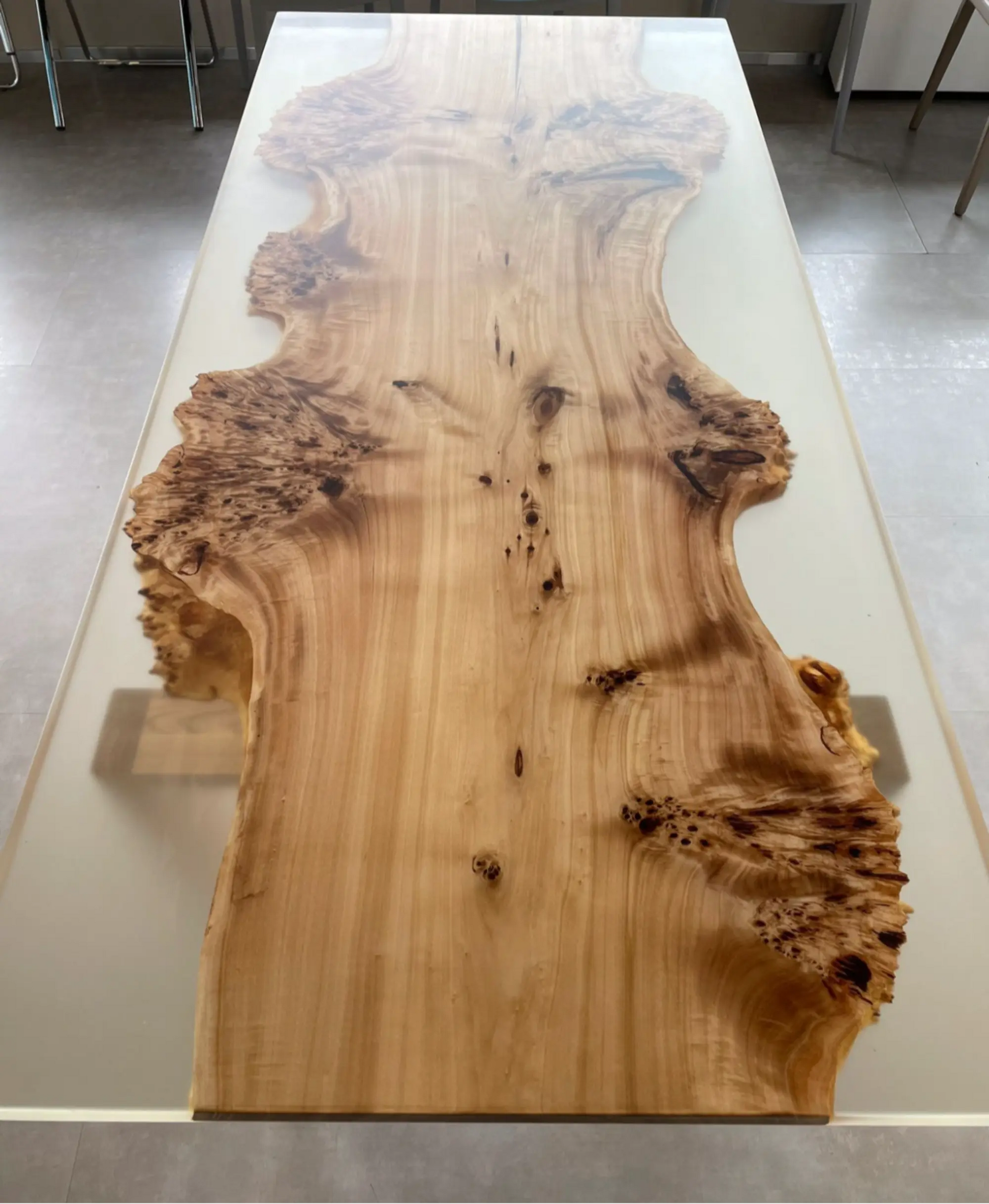 Een houten tafel met een groot blad met natuurlijke randen, waardoor de nerf en textuur van het hout goed tot hun recht komen. De tafel staat op een lichte ondergrond met stoelen op de achtergrond.
