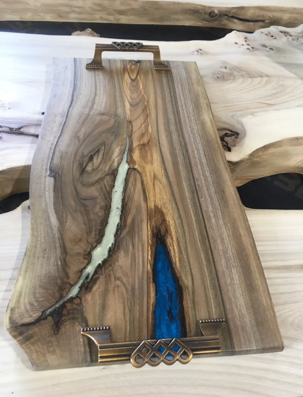 Een gepolijste houten serveerplank met vloeiende natuurlijke nerven en blauwe harsinleg. De plank heeft sierlijke bronzen handgrepen en combineert rustieke en elegante elementen.
