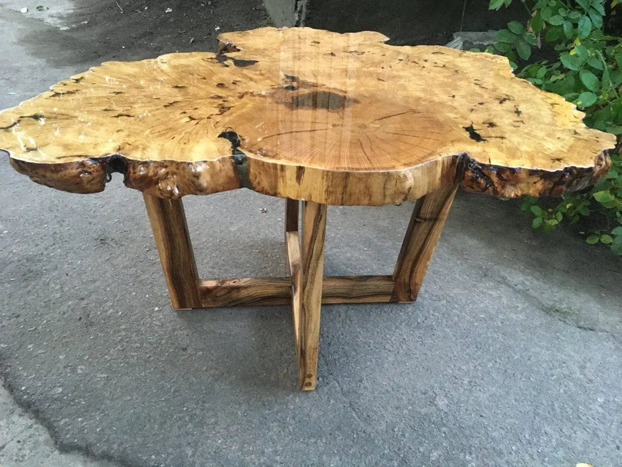 Gepolijste houten tafel met een natuurlijke, onregelmatige rand en unieke nerfpatronen. De tafel staat op stevige houten poten en staat op een betonnen ondergrond.