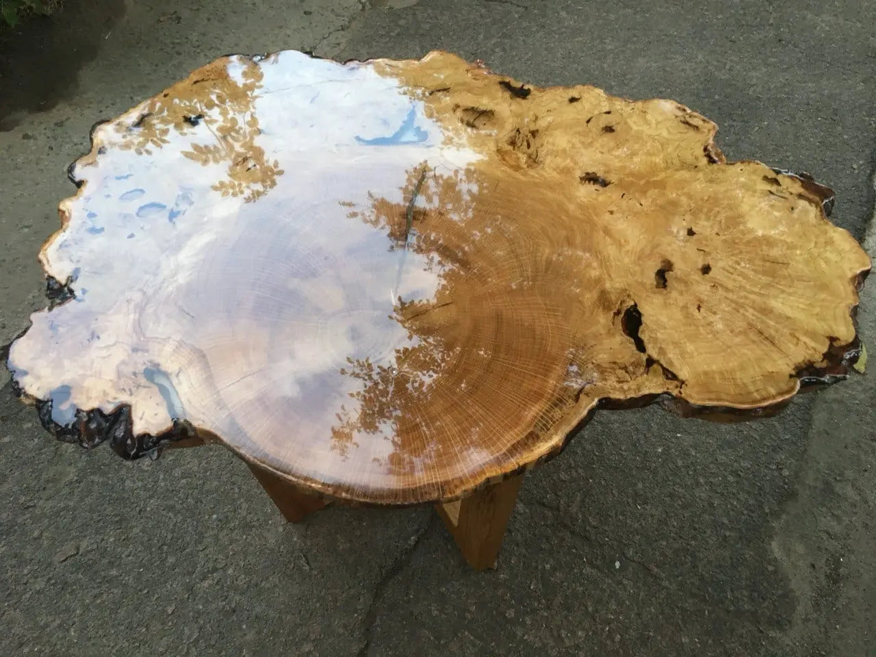 Een gepolijste houten tafel met een ronde, onregelmatige rand, met rijke nerfpatronen en weerspiegelingen van bomen en de lucht, wat een natuurlijke, serene sfeer creëert.