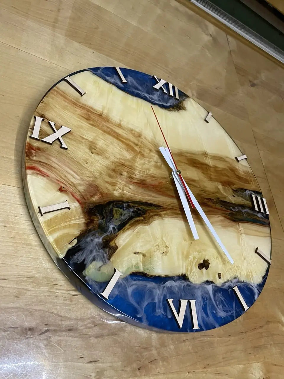 Ronde houten wandklok met Romeinse cijfers en levendig blauwe details van epoxyhars, met wit-rode wijzers. Handgemaakte epoxyklok met rustiek eikenhouten design – perfect voor woninginrichting.