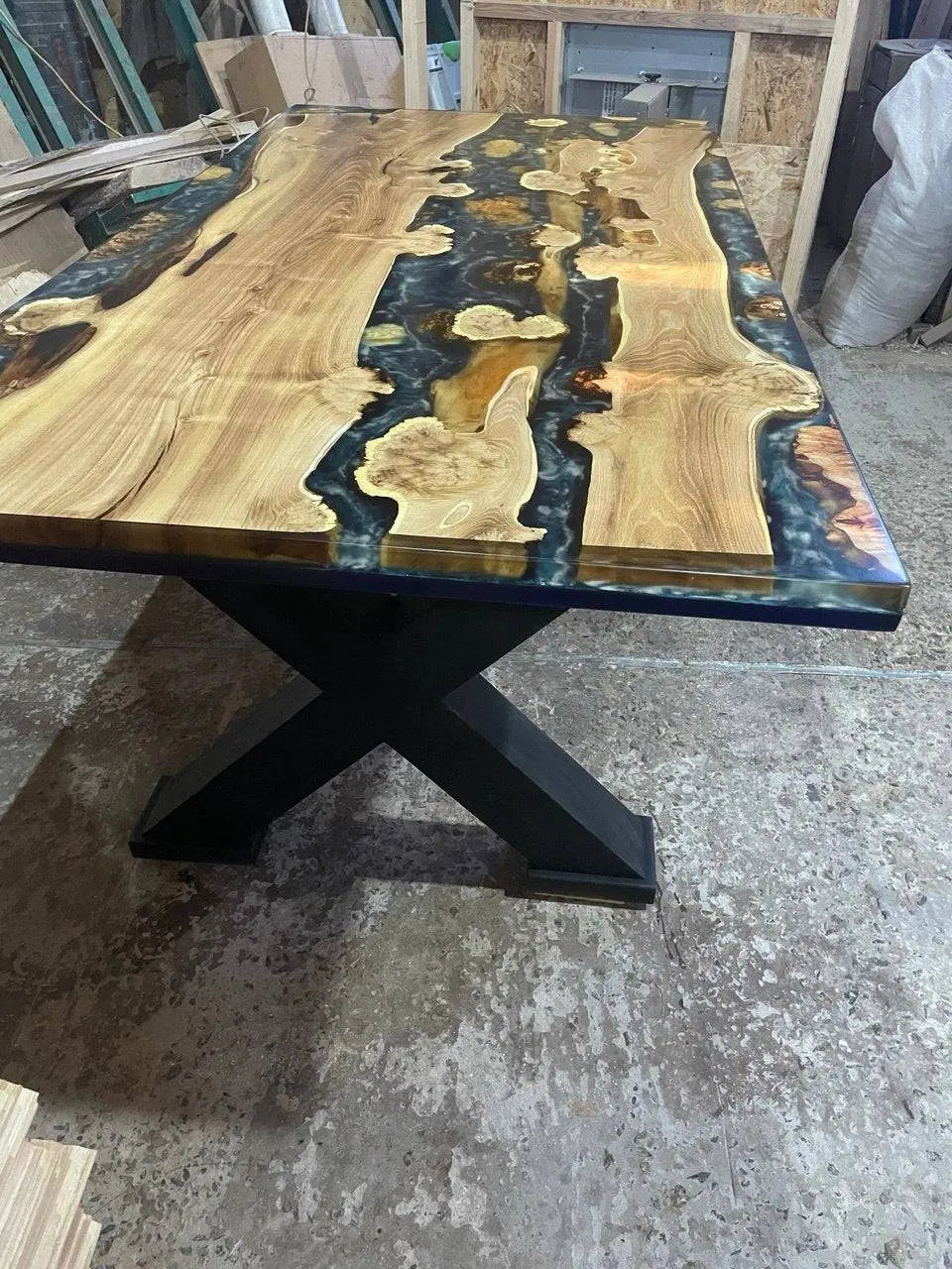 Sideview of smooth wooden table with swirling patterns of polished black resin, resembling a sea, creating a rustic yet elegant atmosphere. Crossed-legs shape of the legs.