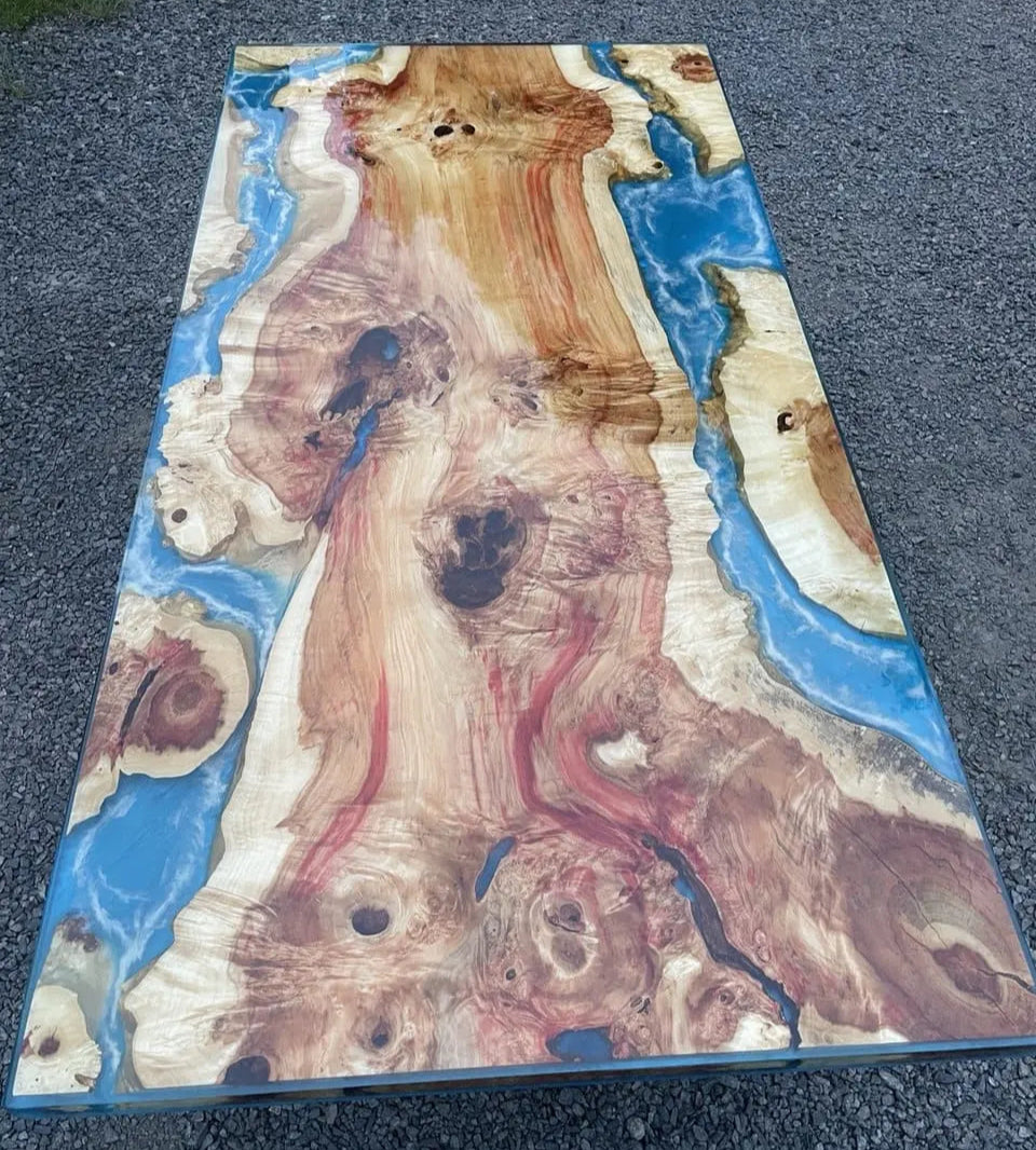 Een rechthoekig tafelblad met een levendig epoxyharsontwerp, waarin wervelende patronen van natuurlijk hout en helderblauw samenkomen. De setting is een buitenruimte met grind, wat een opvallend contrast creëert.