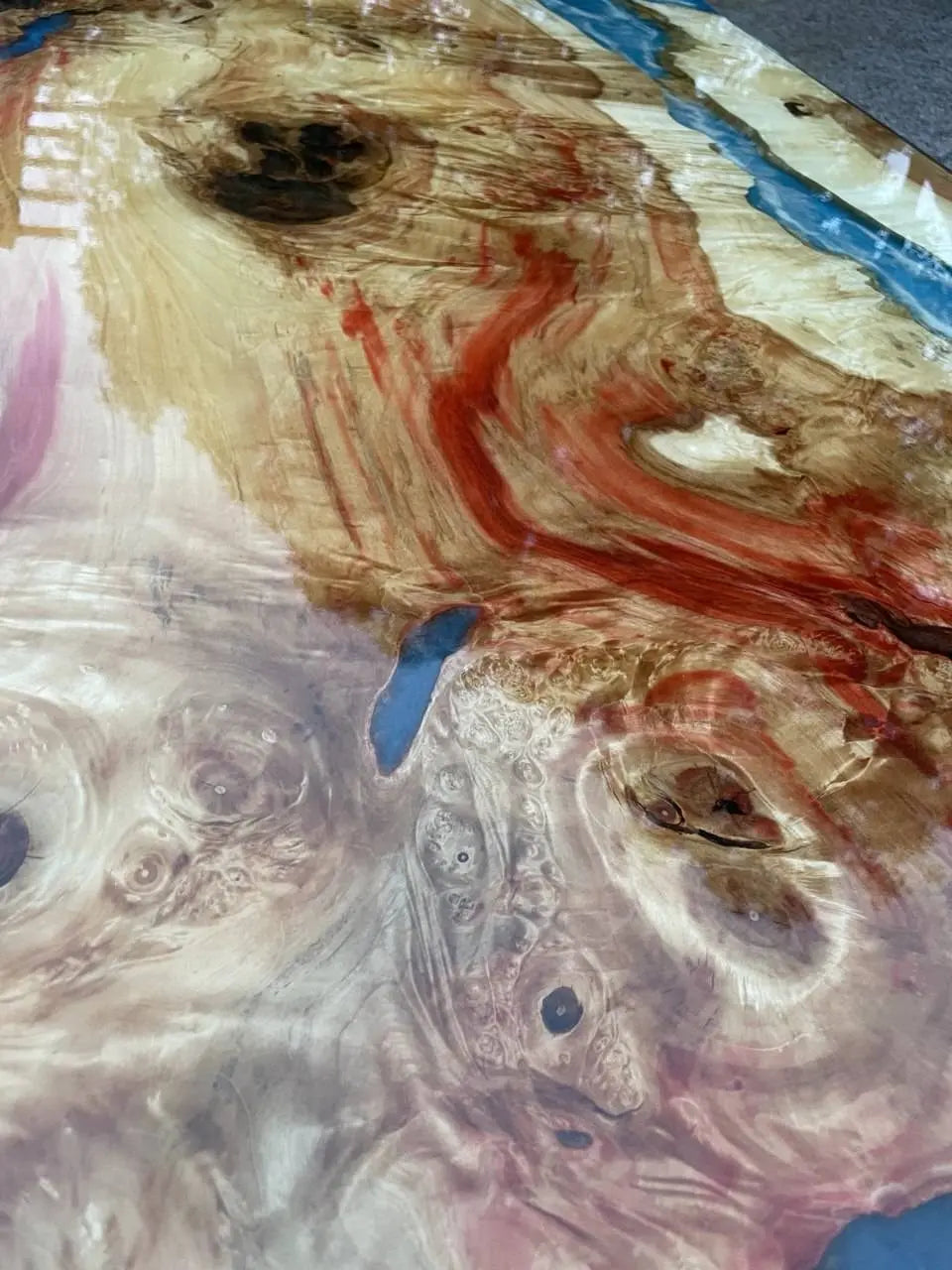 Close-up van een gepolijst houten oppervlak met wervelende rode, beige en bruine patronen. Blauwe accenten zorgen voor contrast en creëren een levendig, abstract ontwerp.