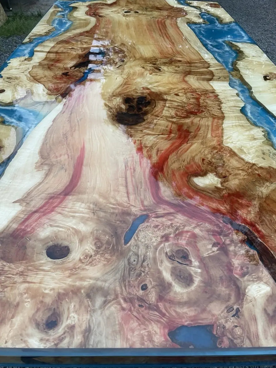 Houten en epoxy tafel met wervelende houtnerf in rood- en bruintinten, doorkruist door levendig blauwe epoxy die doet denken aan een rivier. Het glanzende oppervlak benadrukt de verfijnde texturen en patronen, wat zorgt voor een natuurlijk en artistiek gevoel. De tafel staat buiten op een grindoppervlak.