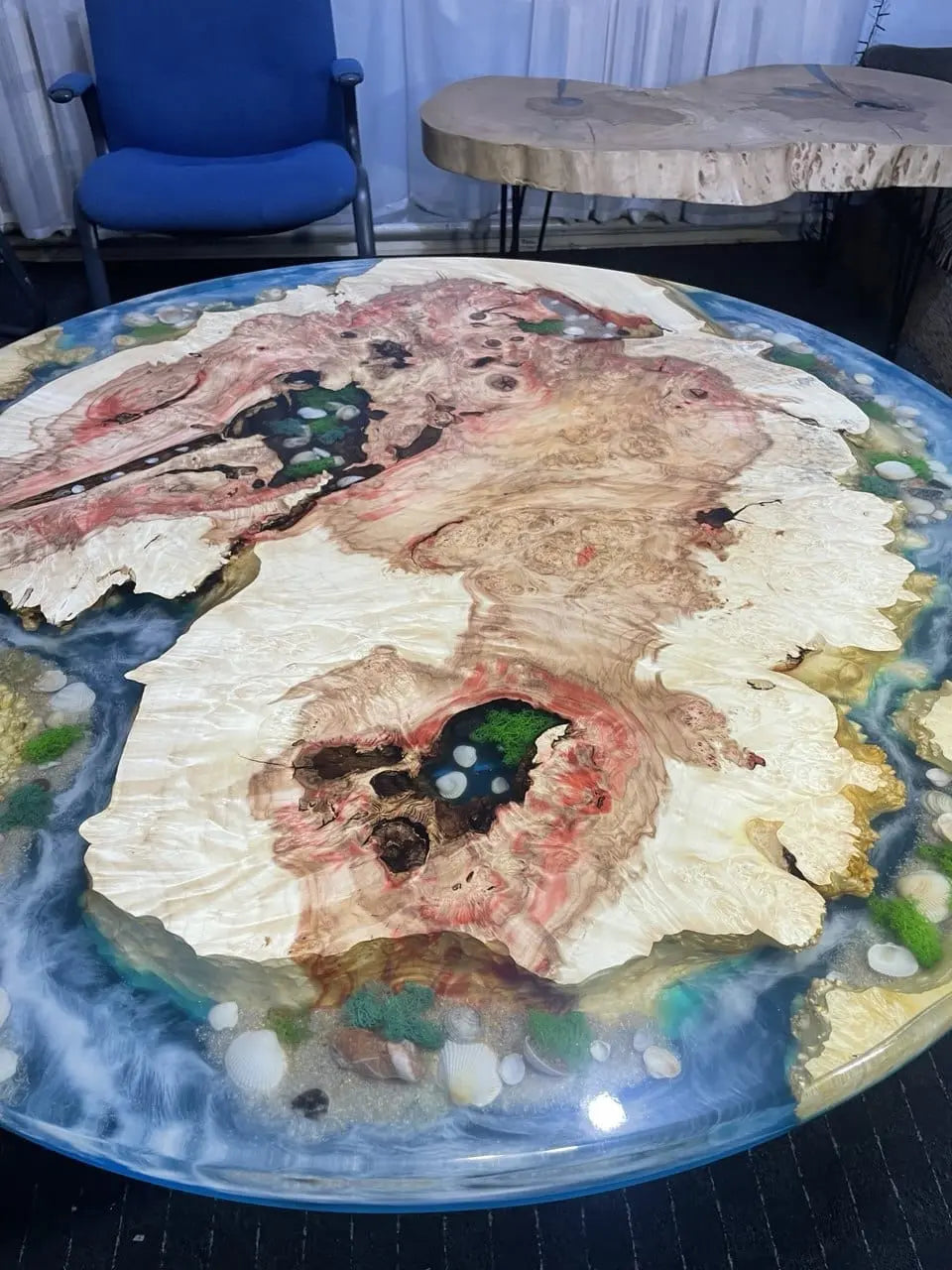 Artistieke ronde houten tafel met wervelende patronen van licht en donker hout. Levendige blauwe epoxy stroomt tussen houteneilanden met ingelegde groene mos en schelpen, wat zorgt voor een organische, kustachtige uitstraling.
