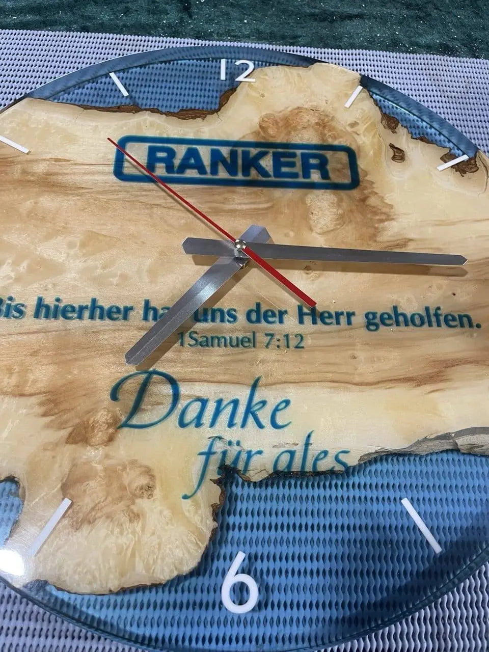 Houten klok met een natuurlijke textuur, met witte cijfers en zilveren wijzers. Een opvallende rode secondewijzer. De tekst bevat een religieus vers en een dankwoord in het Duits.
