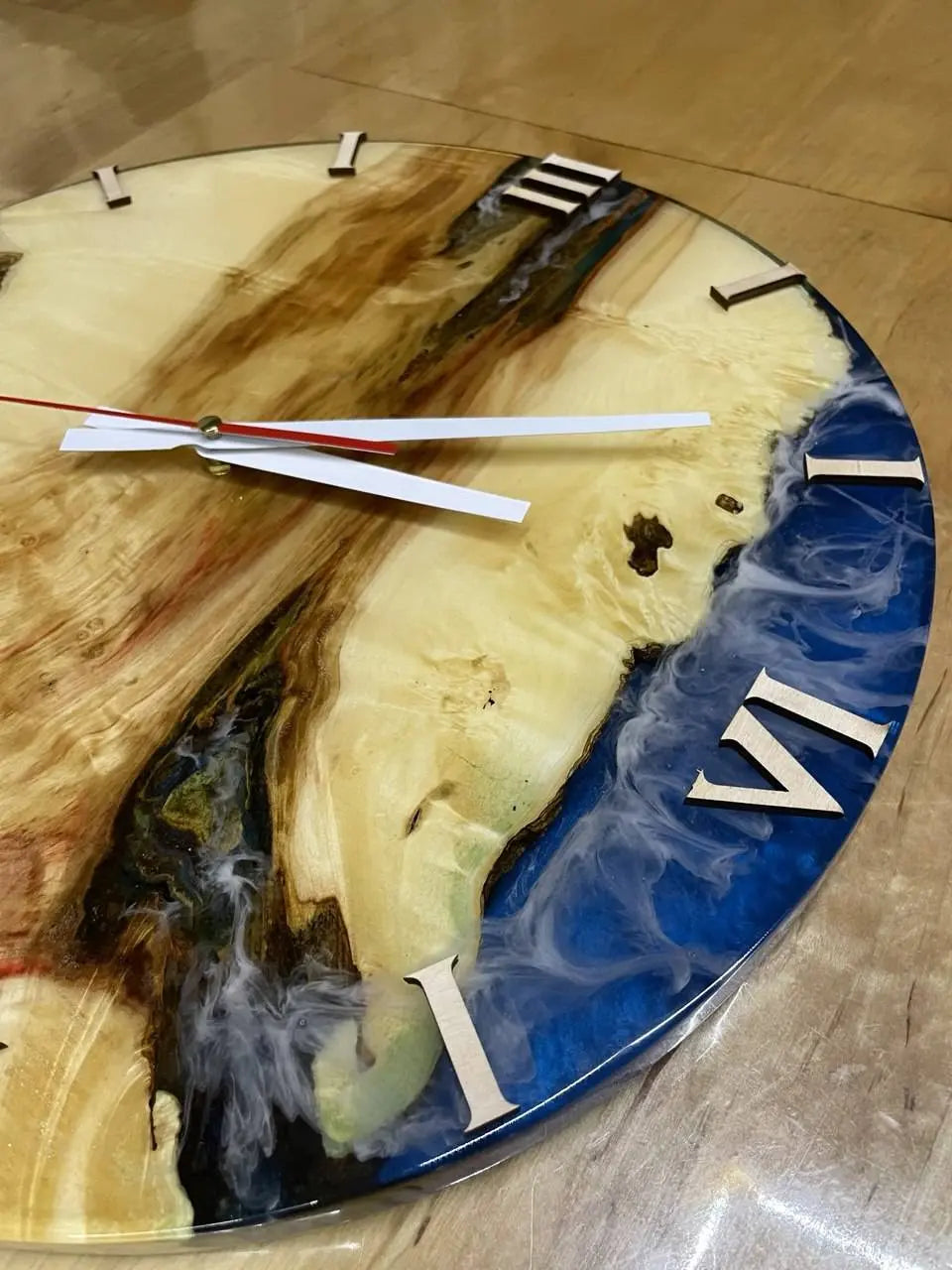 A round wall clock with a resin and wood design. Blue and white swirls blend with natural wood tones. Roman numerals and sleek silver hands.