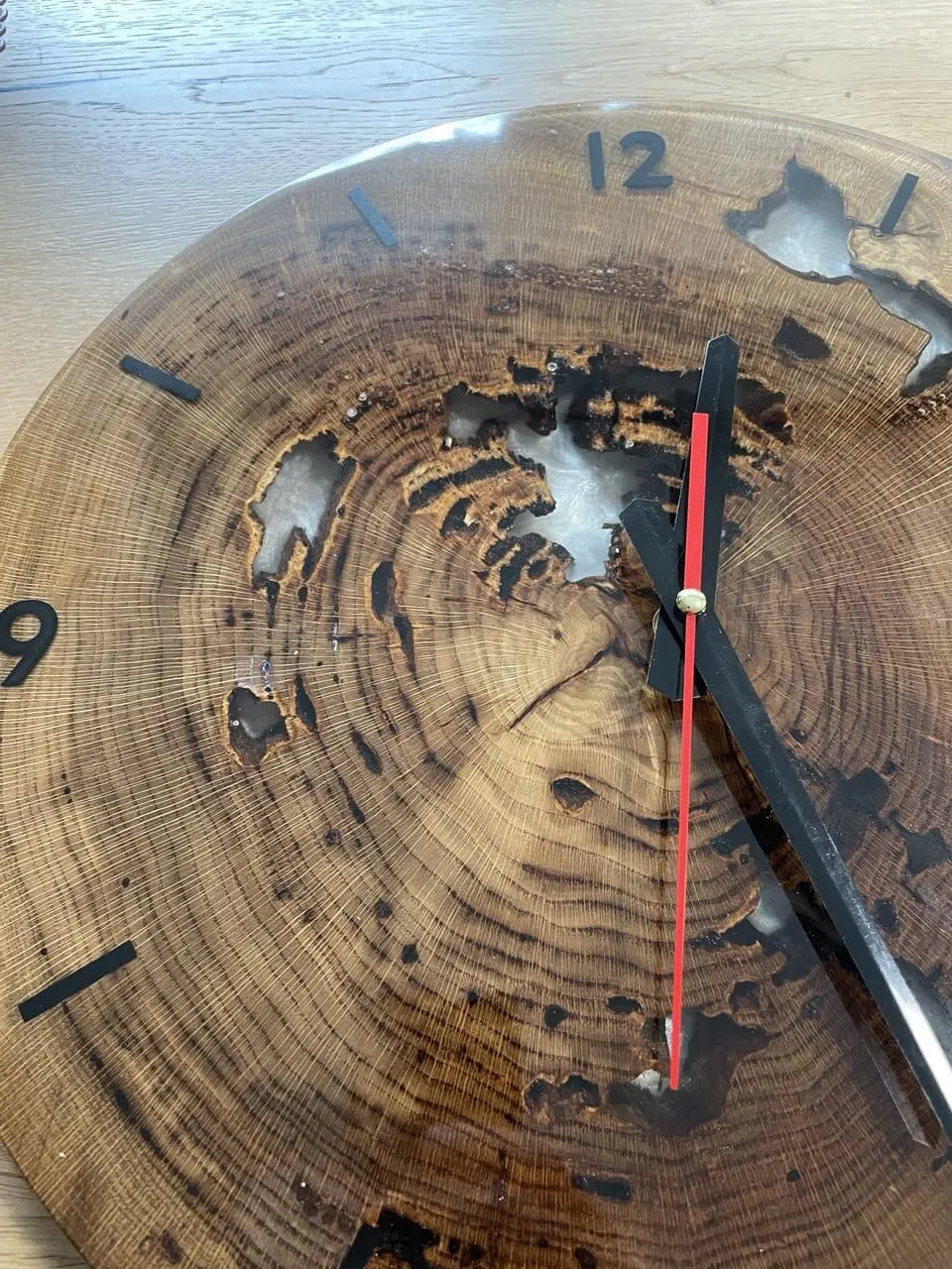 Close-up van een rustieke wandklok gemaakt van een boomstam, met natuurlijke patronen en holtes. Zwarte uurmarkeringen en wijzers met een rode secondewijzer.