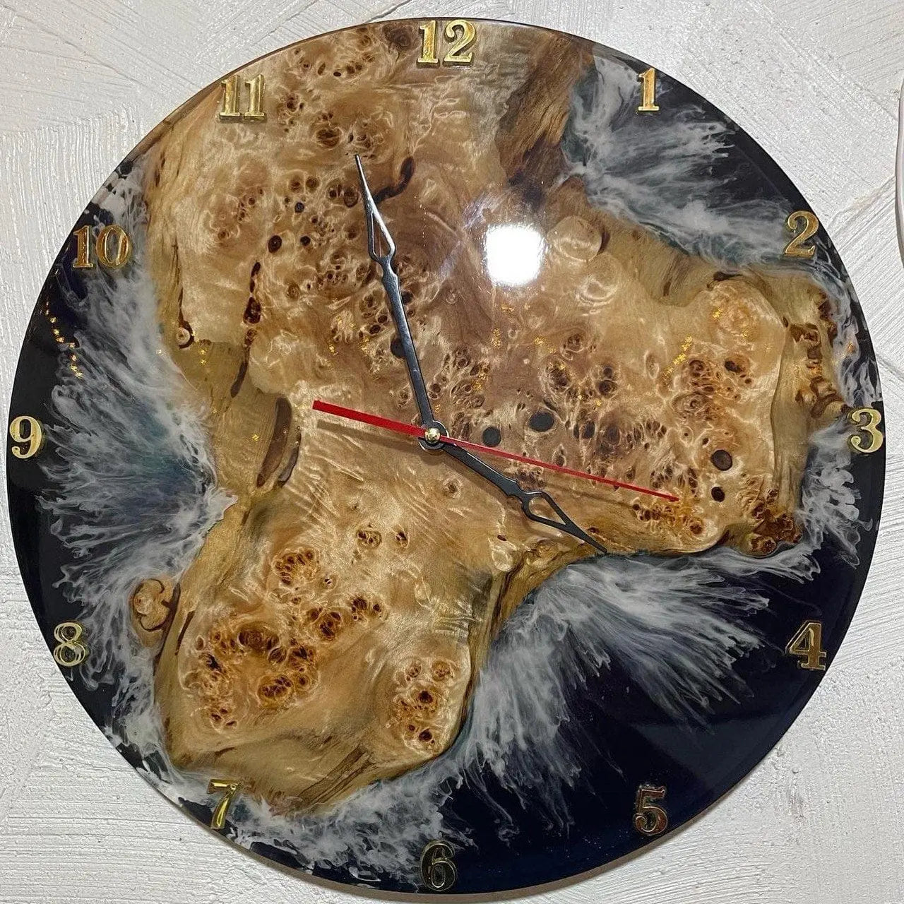 Ronde wandklok met een hout- en harsontwerp, met gouden numerieke uurmarkeringen en drie zwart-rode wijzers. De houtnerf en het hars creëren een natuurlijk, abstract patroon.
