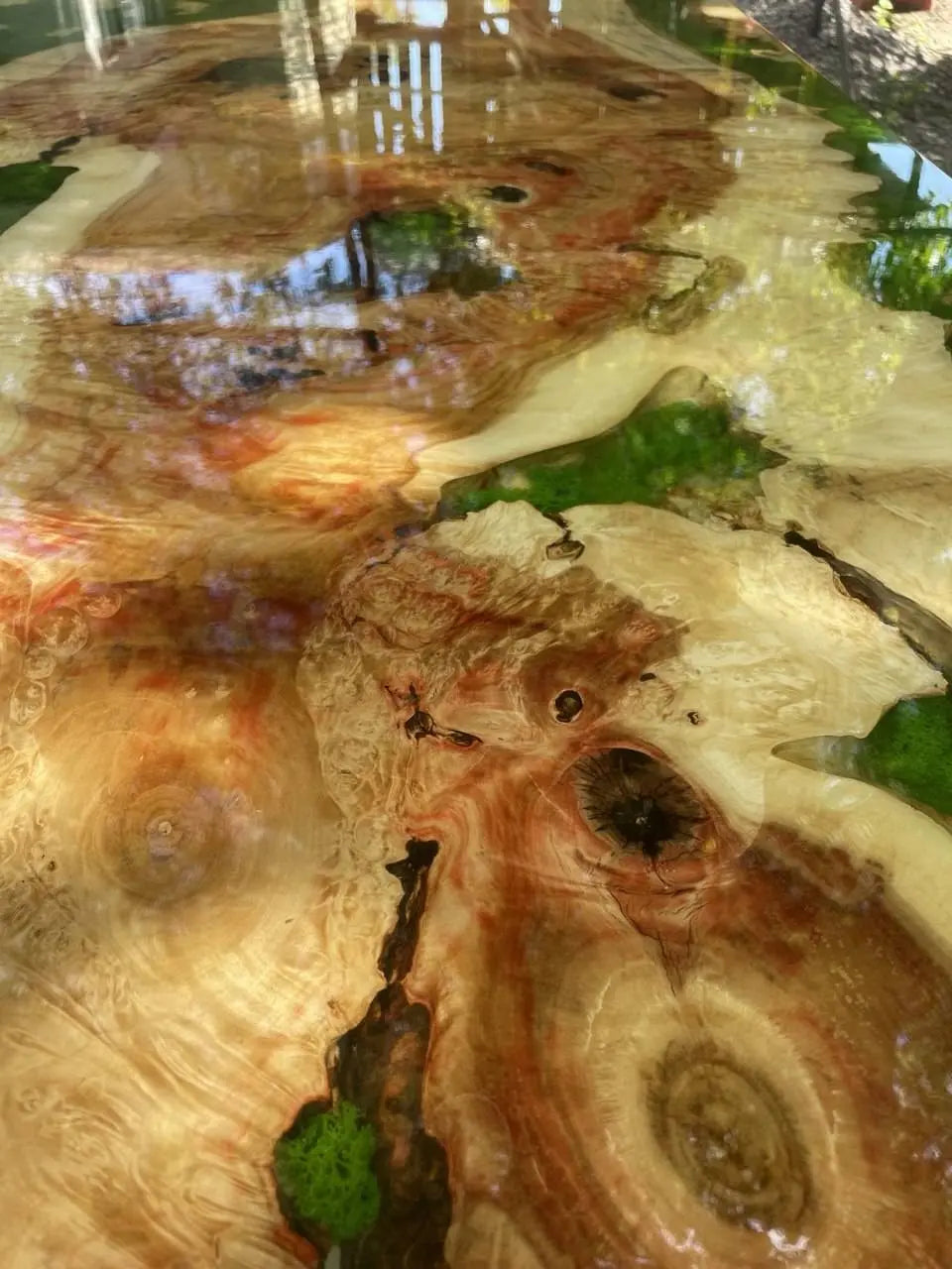 Close-up van een houten tafel met epoxyhars, met wervelende nerfpatronen in amber, bruin en groen. Het oppervlak is glanzend en weerspiegelt licht en de omgeving.