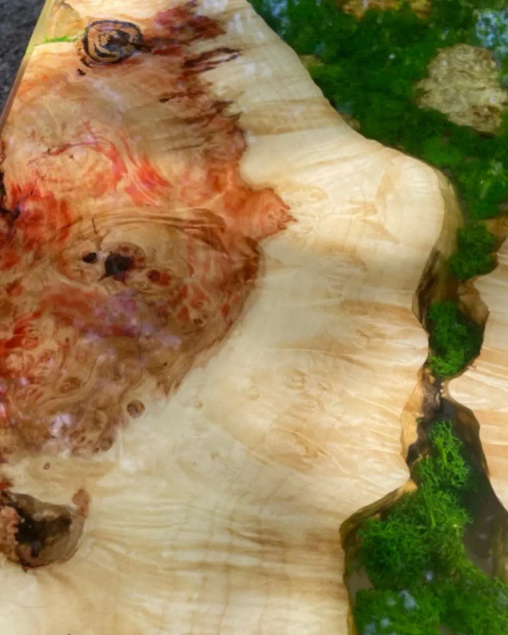 Close-up van een glanzende houten tafel met levendig groen mos ingebed in epoxyhars. Het hout heeft rijke, wervelende nerven met rode en bruine tinten, wat een aards en natuurlijk gevoel creëert.