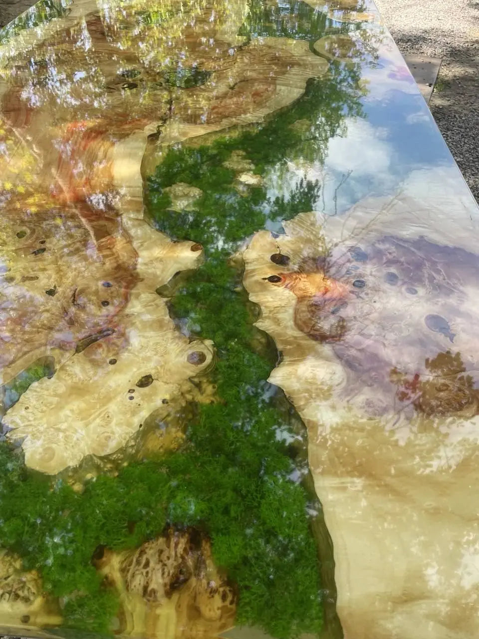 Een glanzend houten oppervlak met wervelende patronen van groen mos en bruine en beige houtnerven, die een lucht met bomen en wolken weerspiegelen, wat een serene, natuurlijke sfeer creëert.
