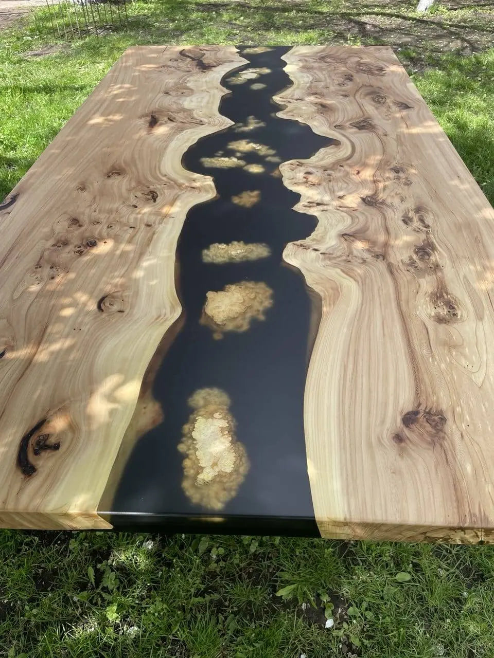 A wooden table with a black resin river flowing through the center, featuring swirling wood grain patterns on each side. The table is placed on lush green grass.