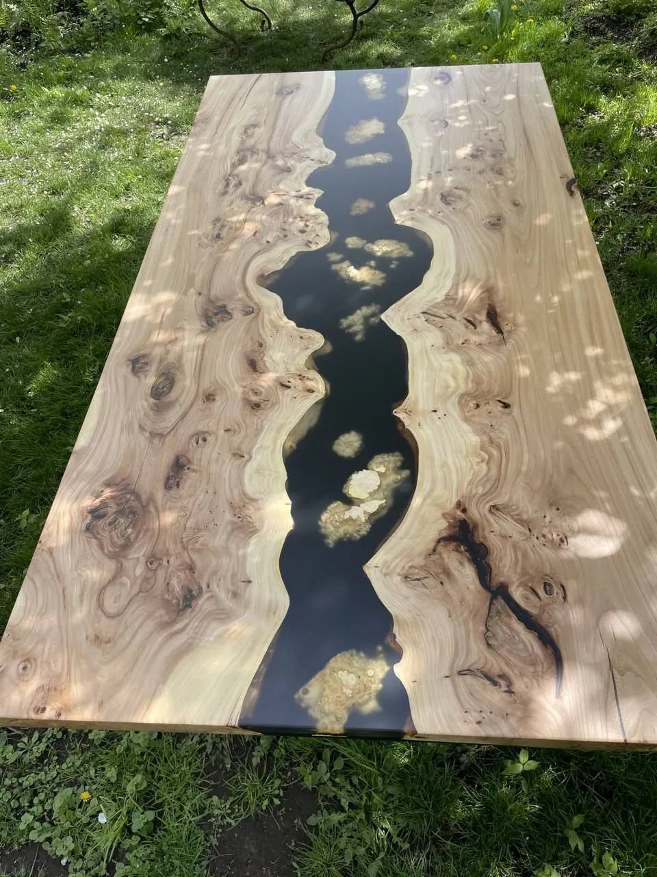 Smooth wooden table outdoors near grass, featuring a river-like black epoxy resin flowing through the center, creating a natural, artistic feel.