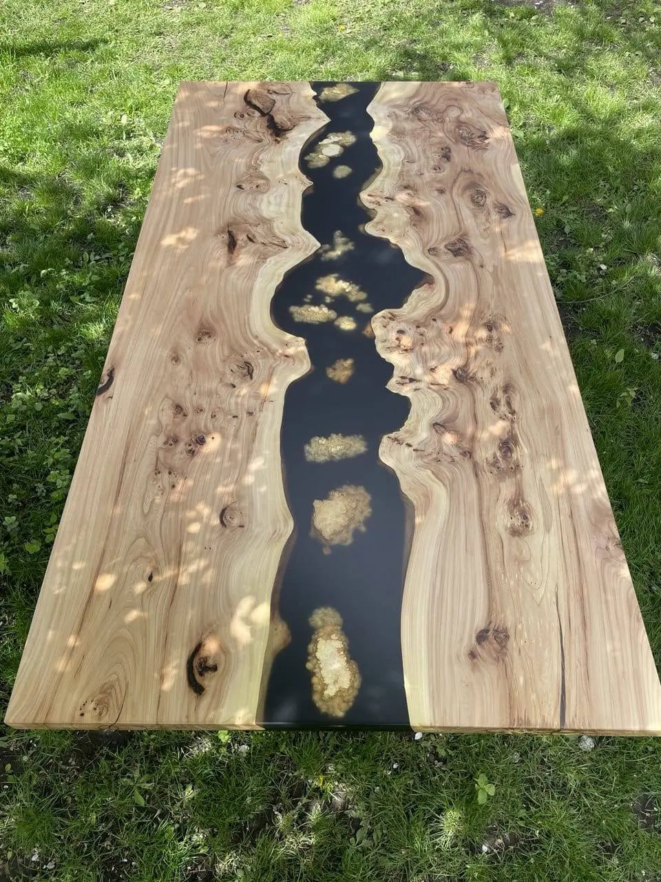 Houten riviertafel voor buiten, met een rivier van zwarte hars die door licht hout met wervelende patronen en knopen stroomt. De tafel staat op groen gras.