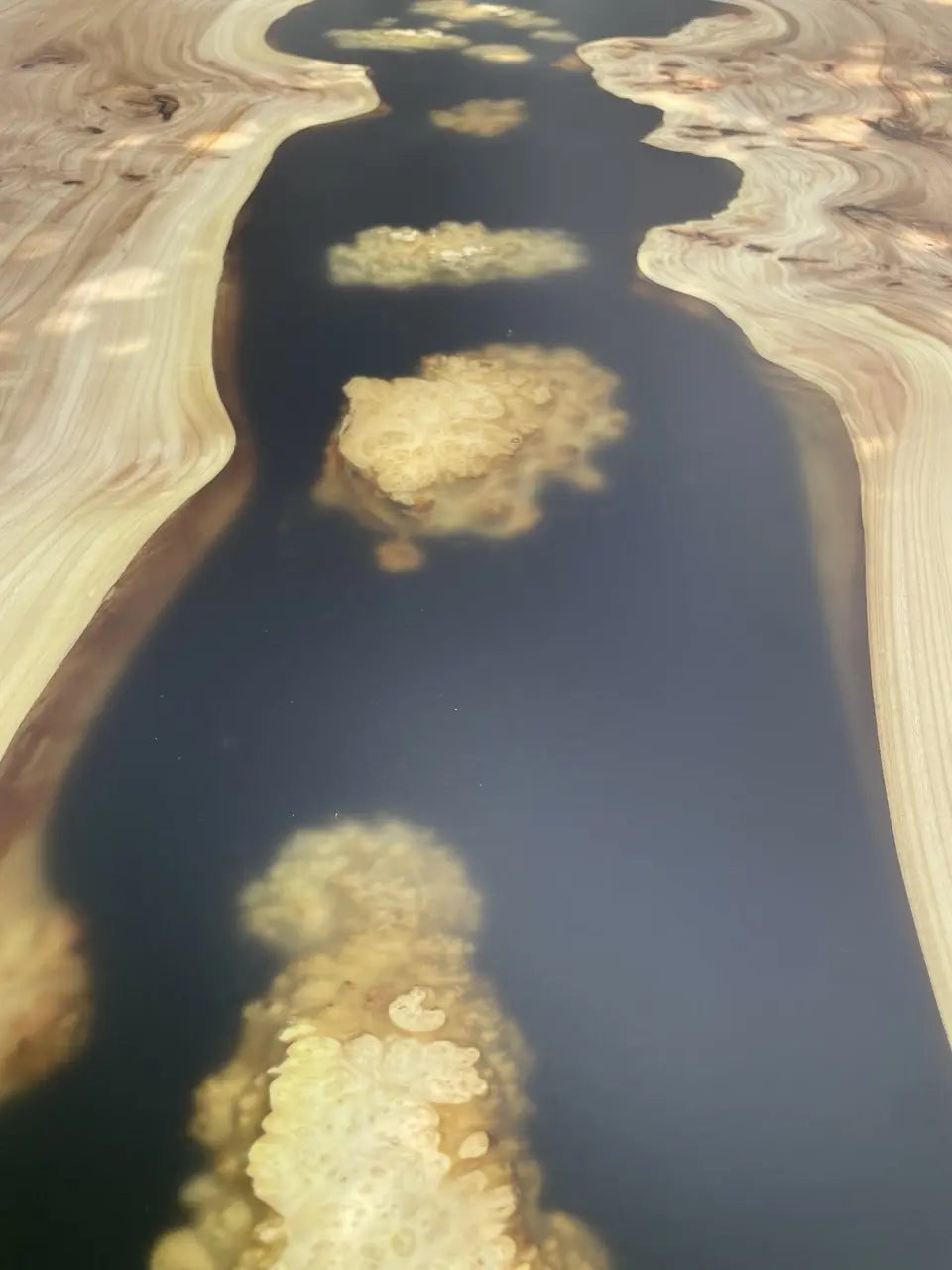 A polished wooden surface with a dark, glossy resin river running through the center, bordered by light, wavy wood grain, creating a natural and elegant look.