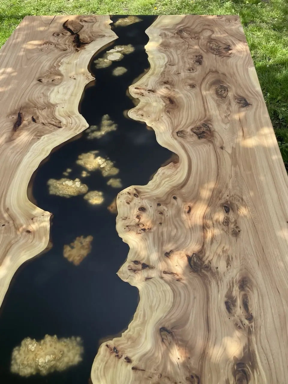 Wooden table with a resin river design. Smooth edges of light and dark wood contrast with a black resin center. Sunlight dapples the surface gently.