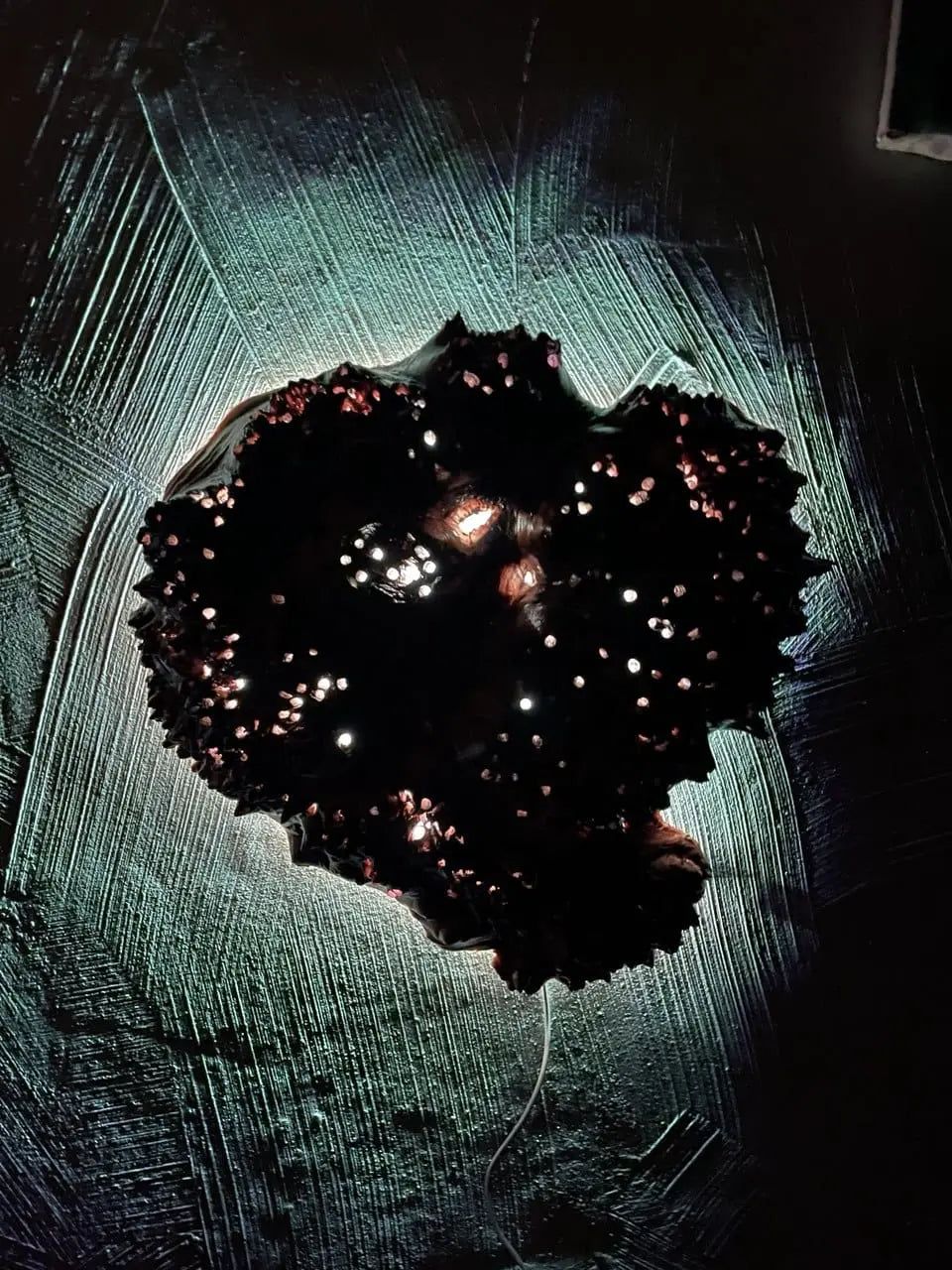 Abstract kunstwerk met een getextureerde, donkere cluster met kleine, gloeiende witte en rode lichtjes. Een blauwgroene halo en lineaire patronen omringen de cluster op een zwarte achtergrond.