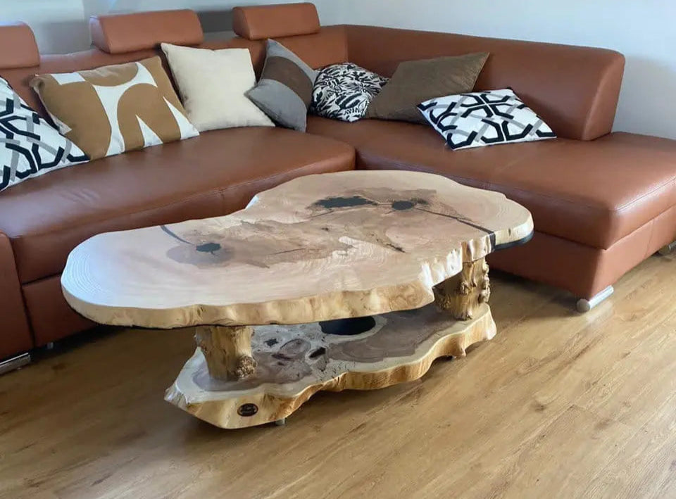 Een houten salontafel met natuurlijke randen en donkere markeringen staat op een lichte houten vloer. Erachter liggen een bruine hoekbank met effen kussens met een patroon.