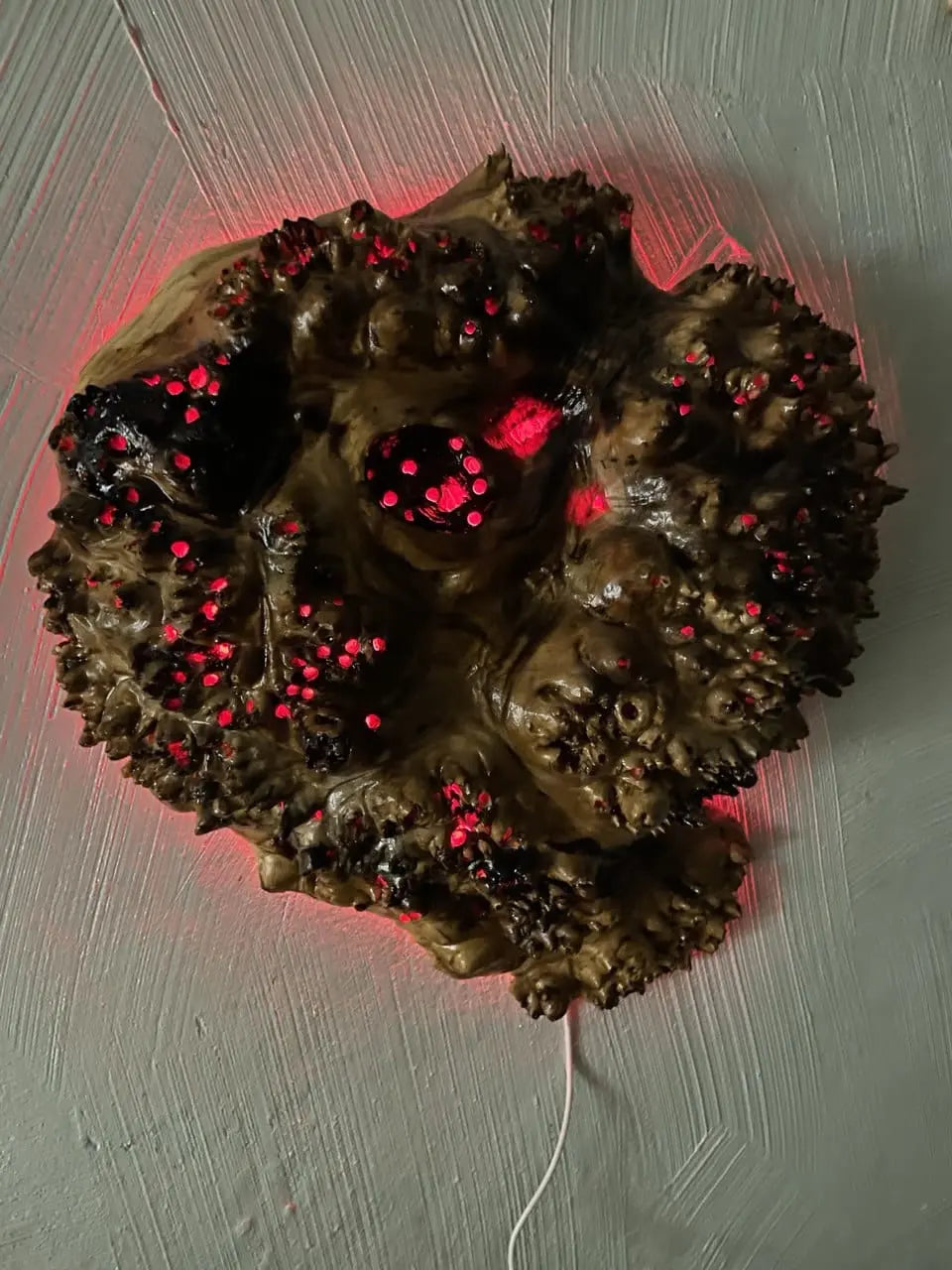 Een organisch, rond beeld met een ruwe textuur en oplichtende rode stippen, gemonteerd op een grijze muur, straalt een mysterieuze, sciencefictionachtige sfeer uit.