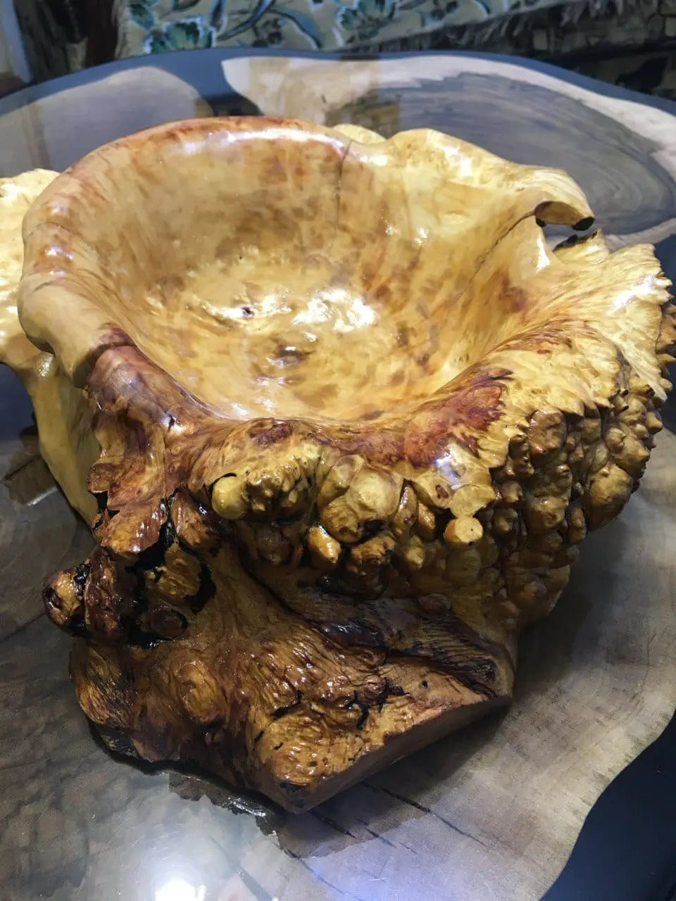 A polished wooden bowl with a smooth, glossy finish and an irregular, natural edge sits on a dark wooden table. It conveys an earthy, rustic feel.