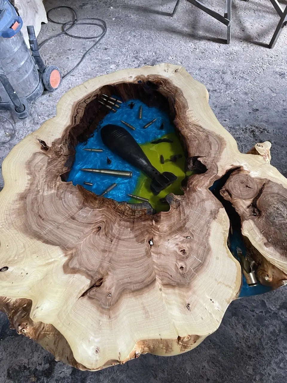 Tafel van natuurlijk hout met in het midden een epoxy-inleg die op water lijkt, met daarop een groot, donker object en verspreide kogels, in een rustieke werkplaats.