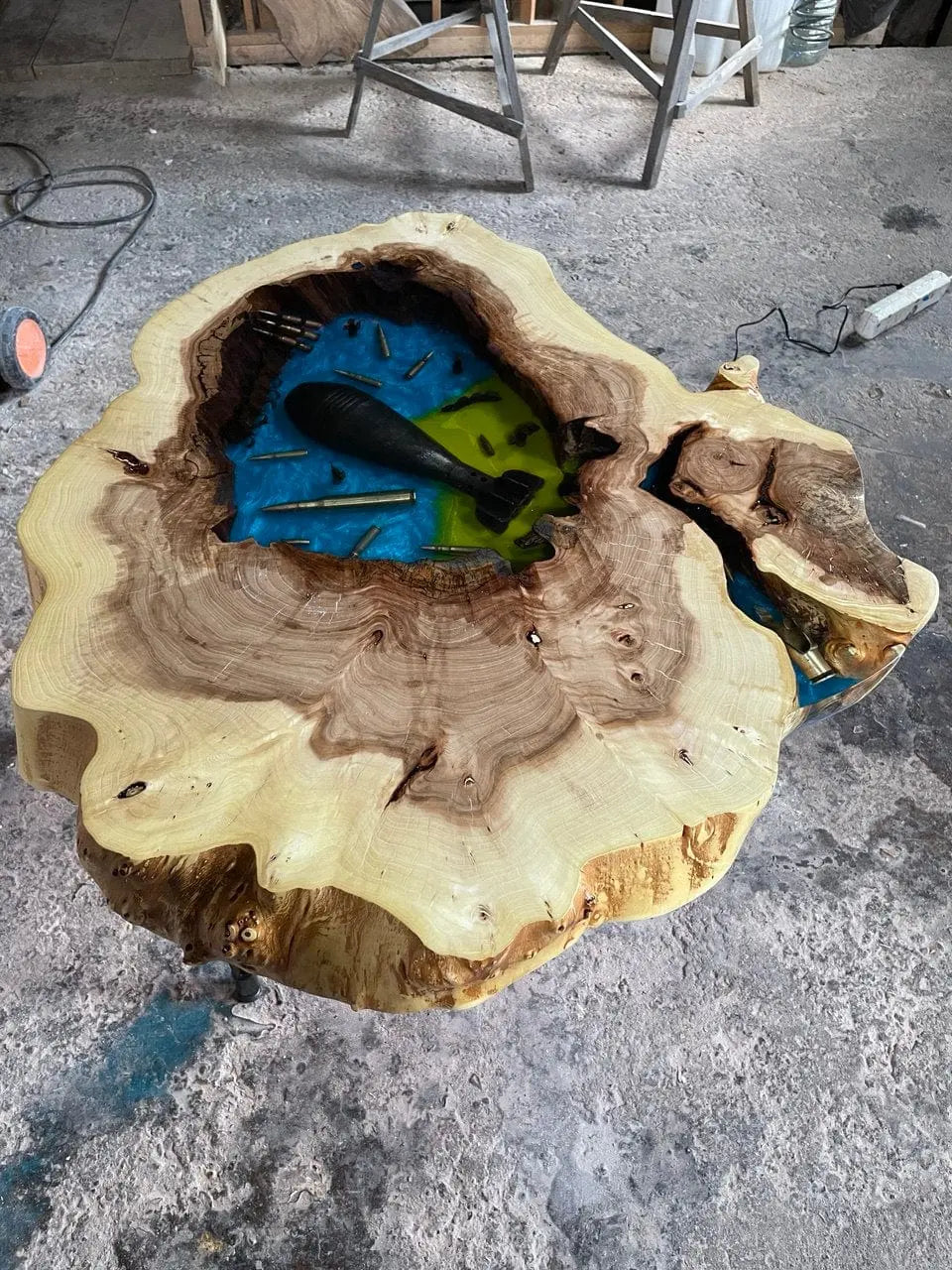 Handgemaakte rustieke houten salontafel met een natuurlijke boomrand en kleurrijke epoxy inleg, gevuld met echte kogelhulzen en bomscherven. Een uniek pronkstuk op de vloer van een werkplaats.