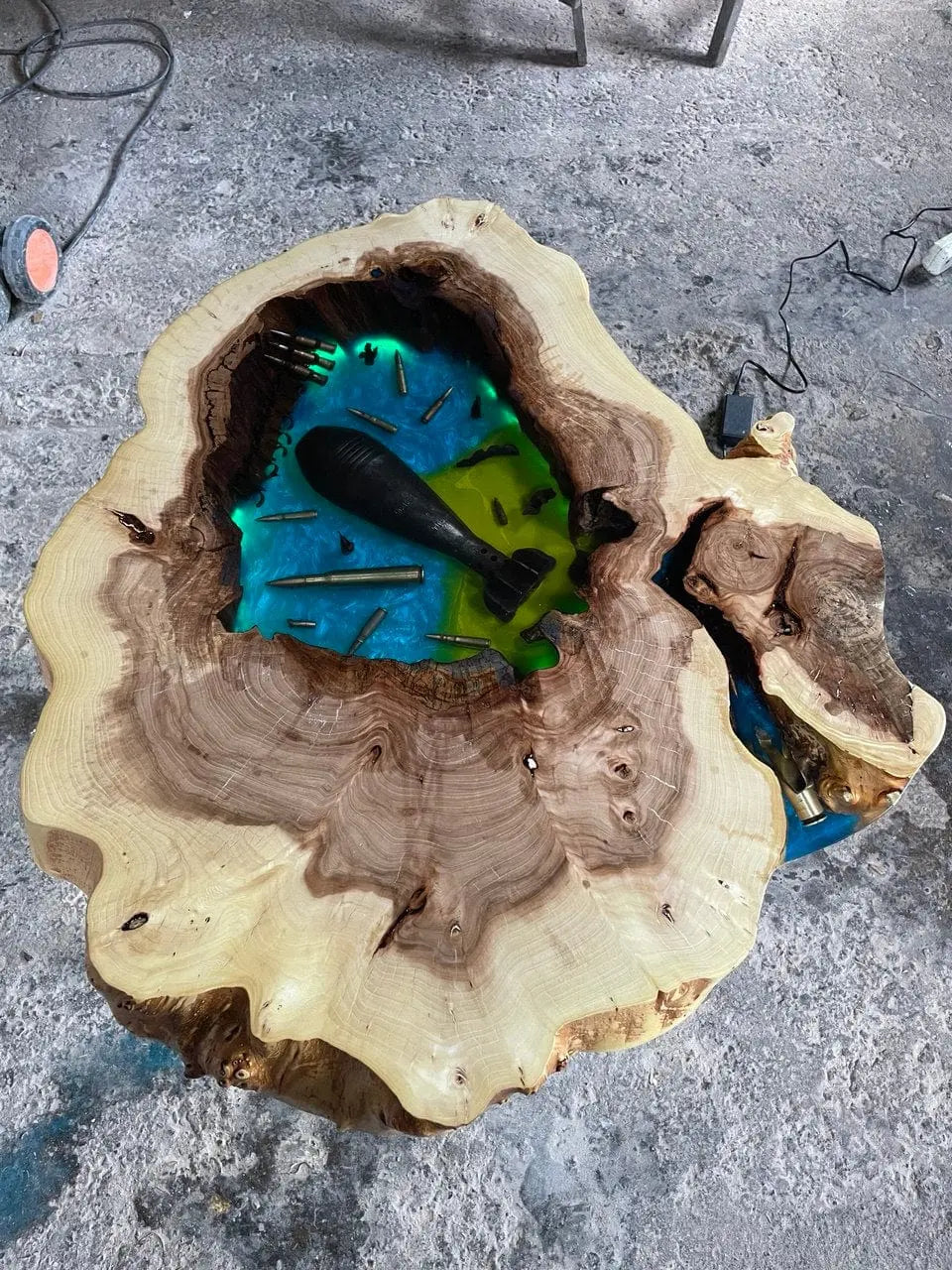 Het tafelblad is handgesneden uit massief iepenhout en afgewerkt voor een natuurlijke, robuuste uitstraling. Eén kant van de tafel is indrukwekkend gevuld met echte kogelhulzen, een granaat en diepblauwe epoxyhars.