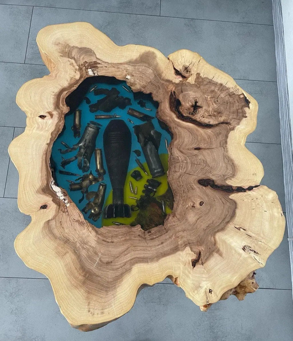 Houten tafel met een natuurlijke, onregelmatige rand, met een opvallend blauw en groen harsen middenstuk met daarin verwerkte kogels en een bomvorm, wat een robuuste, artistieke sfeer uitstraalt.