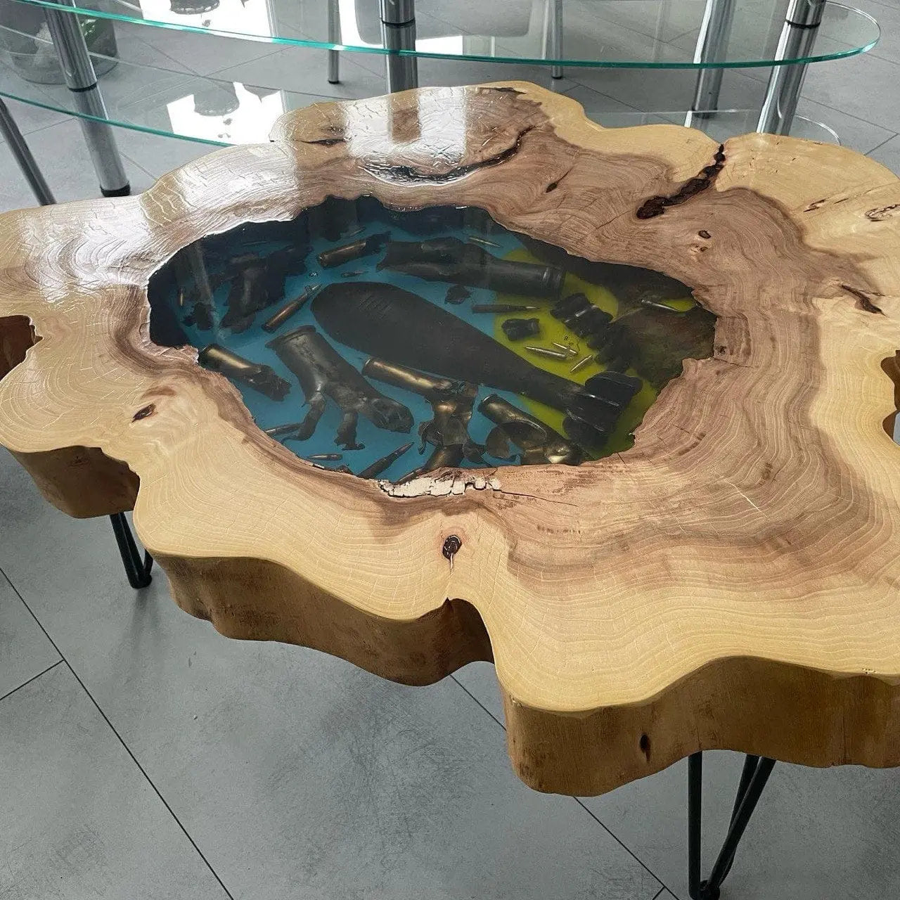 Tafel van natuurlijk hout met een middenstuk van glas-epoxy in blauw- en groentinten en met ingelegde snuisterijen, op zwarte metalen haarspeldpoten, in een kantooromgeving.