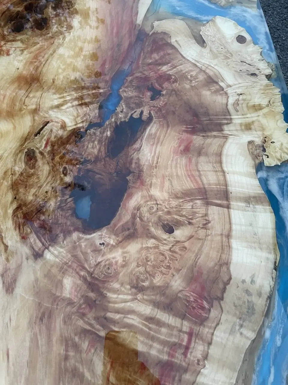 Close-up van een gepolijst houten oppervlak met wervelende nerfpatronen in beige, bruine en rode tinten, tegen een helderblauwe achtergrond, wat een natuurlijke, artistieke uitstraling geeft.