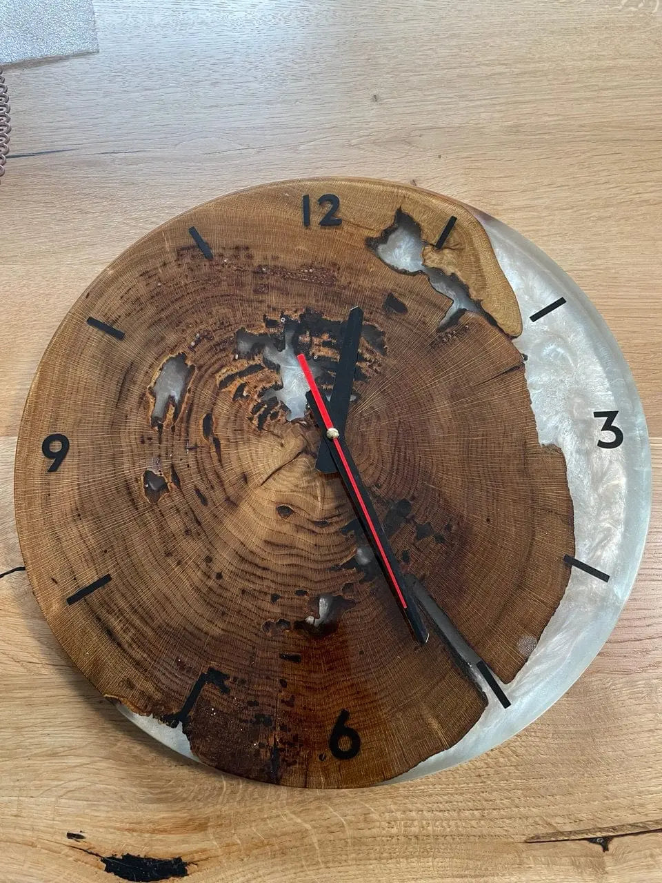 Houten klok met natuurlijke boomringen en harsinleg, zwarte uurmarkeringen en rode wijzers op een licht houten oppervlak. Rustieke, artistieke uitstraling.