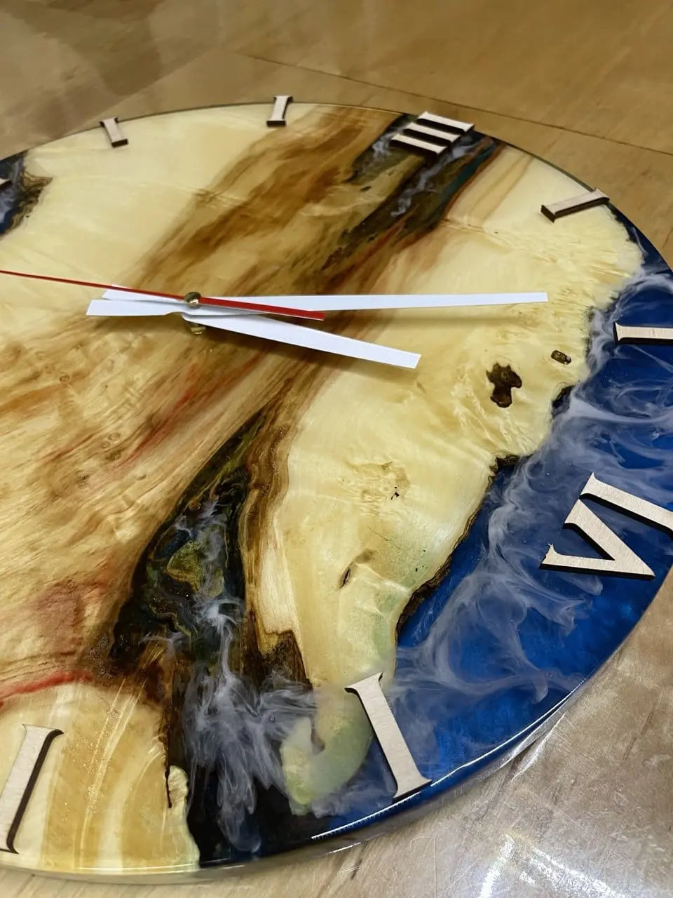 Een ronde houten klok met een glanzende harsafwerking, waarin natuurlijke houttinten worden gecombineerd met blauwe en witte spiralen. Romeinse cijfers en strakke witte wijzers.